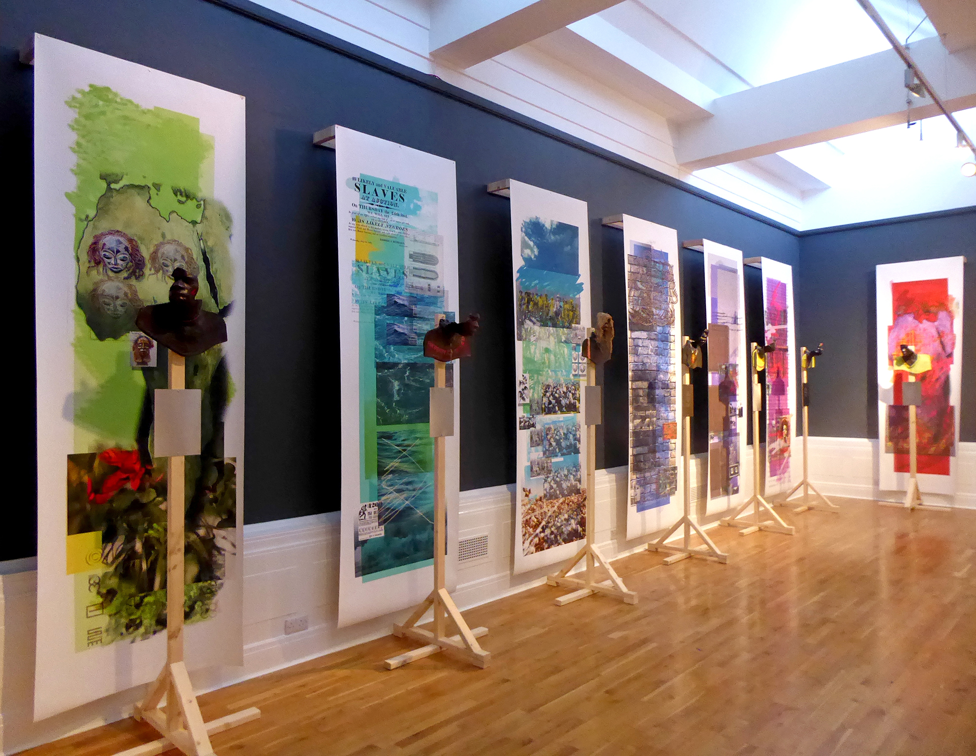 A series of seven hanging, horizontal colourful collages. Each collage has different images from faces to landscapes and brickwork. In front of each collage is a partial sculpture of a head 