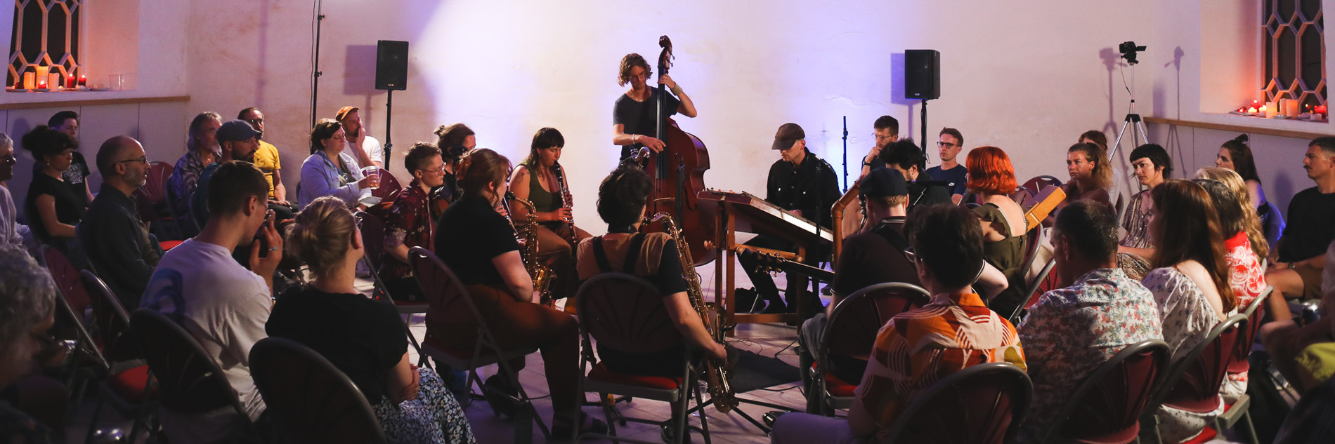 A photograph of a group of adults playing musical instruments. An audience of adults are sitting on chairs surrounding the performers.