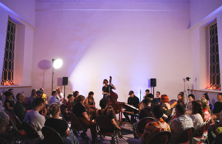 A photograph of a group of adults playing musical instruments. An audience of adults are sitting on chairs surrounding the performers.