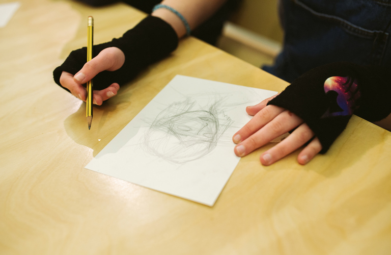 A work in progress pencil drawing of a person's head on a table with someone's hands either side. 