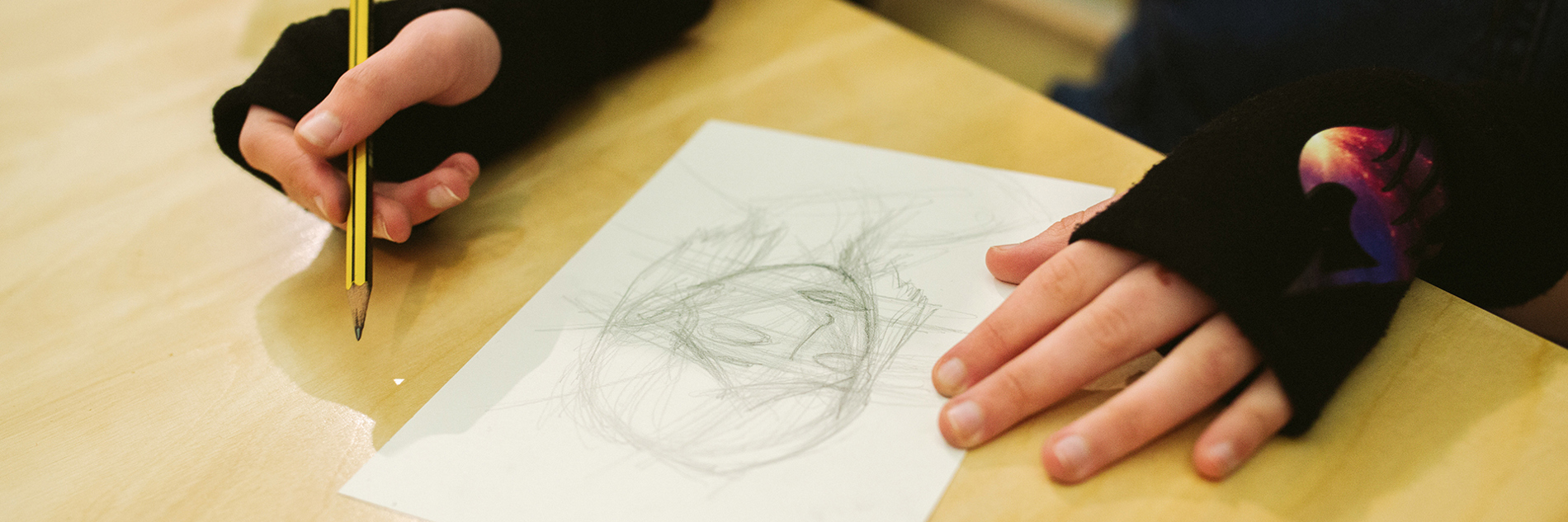 A work in progress pencil drawing of a person's head on a table with someone's hands either side. 