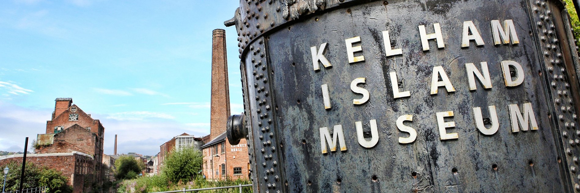 Huge metal industrial bucket with 'Kelham Island Museum' written on. Various buildings including one with a tall chimney are in the background.