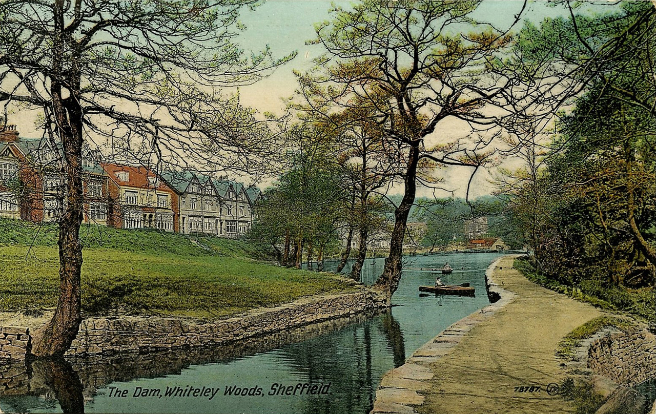 A historic colour photographic postcard image of a pond in a park. On one side of the pond is a footpath and on the other side is a grassy area. In the distance there are a row of houses. 