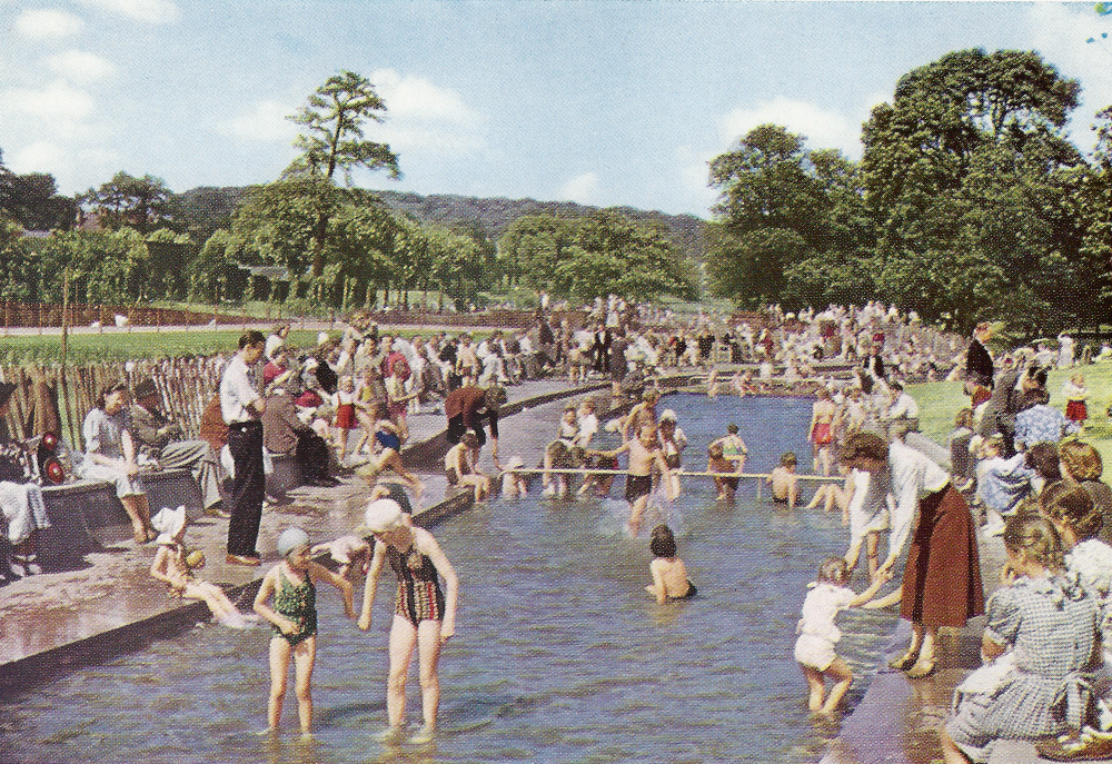 Children in swimsuits play in a large outdoor paddling pool in a park on a sunny day. Adults watch and assist them from the poolside.  In the background is grass and green trees.