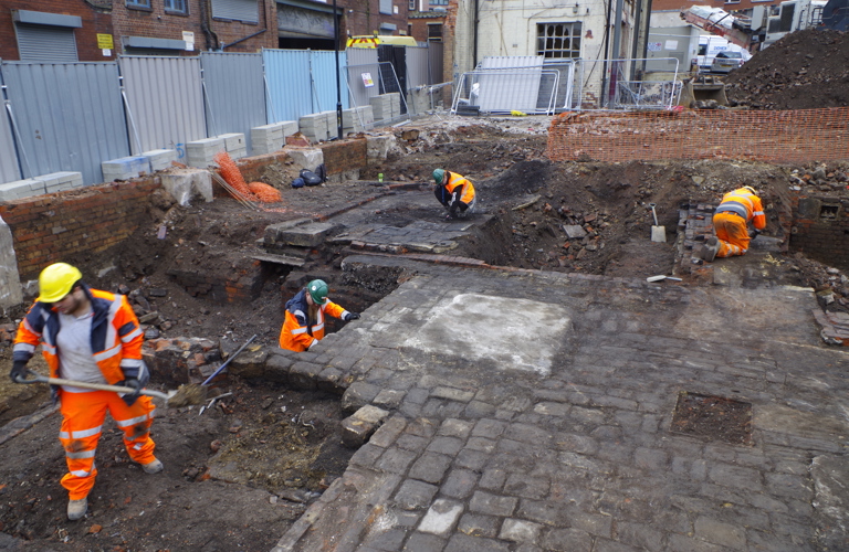 A photograph of four workers in orange high vis and hard hats excavating the Sheffield Castle dig site. There are boards and scaffolding surrounding the site and various stone work paths that the workers are carefully working around.