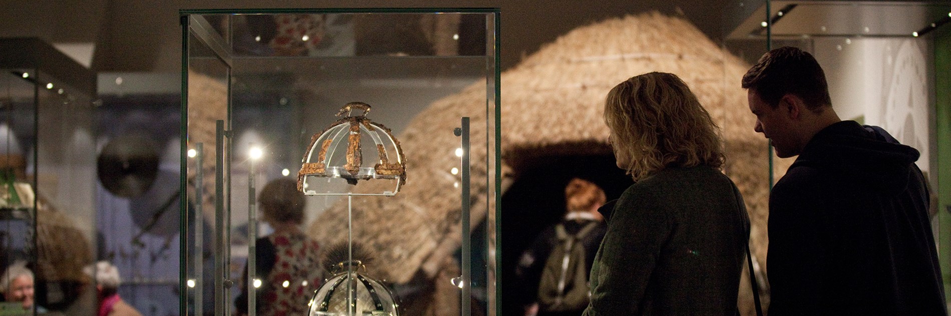 Two adults stood looking at a display case with the remains of a helmet with a boar on top - there is a modern replica below the original. The background is a exhibition area with visitors and other displays including the Iron Age Roundhouse.