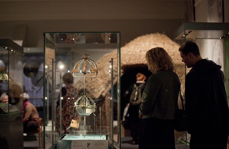 Two adults stood looking at a display case with the remains of a helmet with a boar on top - there is a modern replica below the original. The background is a exhibition area with visitors and other displays including the Iron Age Roundhouse.