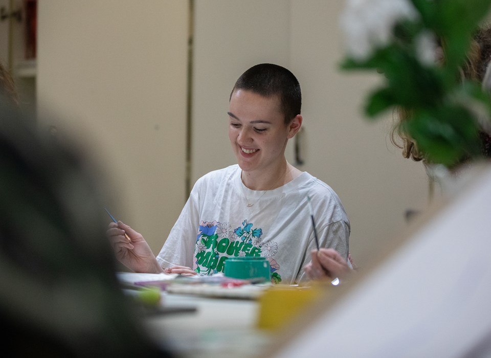A smiling young person with wearing a white t-shirt paints at a crating table.