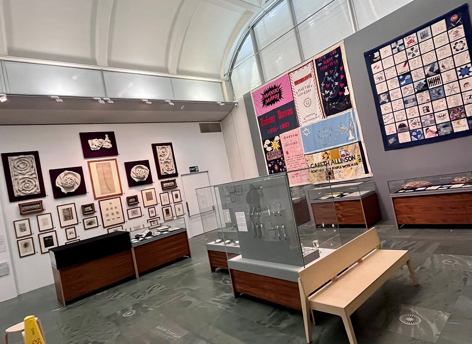 A modern gallery space with a curved ceiling> on the right hand wall are two embroidered patchwork quilts. On the left hand way are a series of works on paper and plaster reliefs. In the foreground are a series of display cases and a modern light wood bench. 