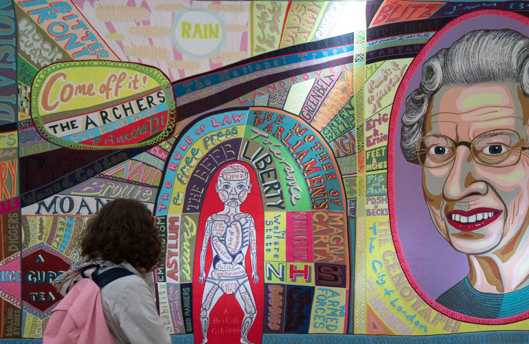 A person looks at a colourful large-scale tapestry which features words and symbols associated with British life, including a large depiction of Queen Elizabeth II