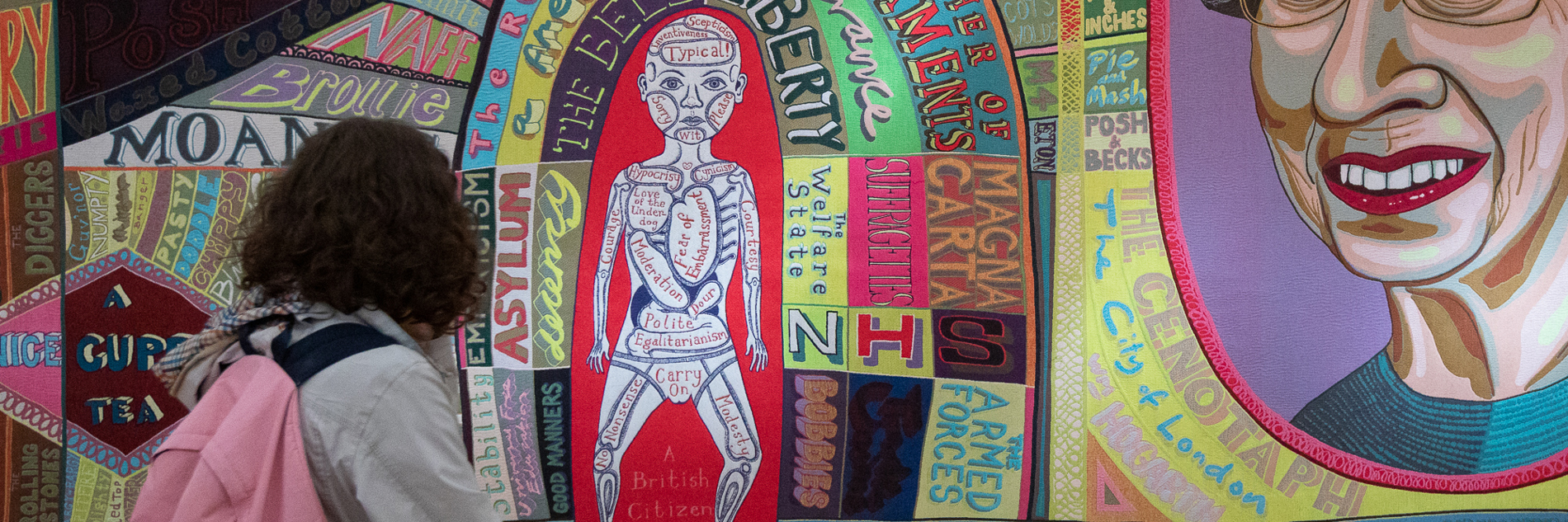 A person looks at a colourful large-scale tapestry which features words and symbols associated with British life, including a large depiction of Queen Elizabeth II