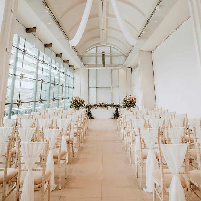A contemporary white room with a vaulted ceiling and floor to ceiling windows on one side dressed for a wedding. Fabric has been hung from the centre of the ceiling and used to decorate rows of chairs. At far end of the room are three floral arrangements.   