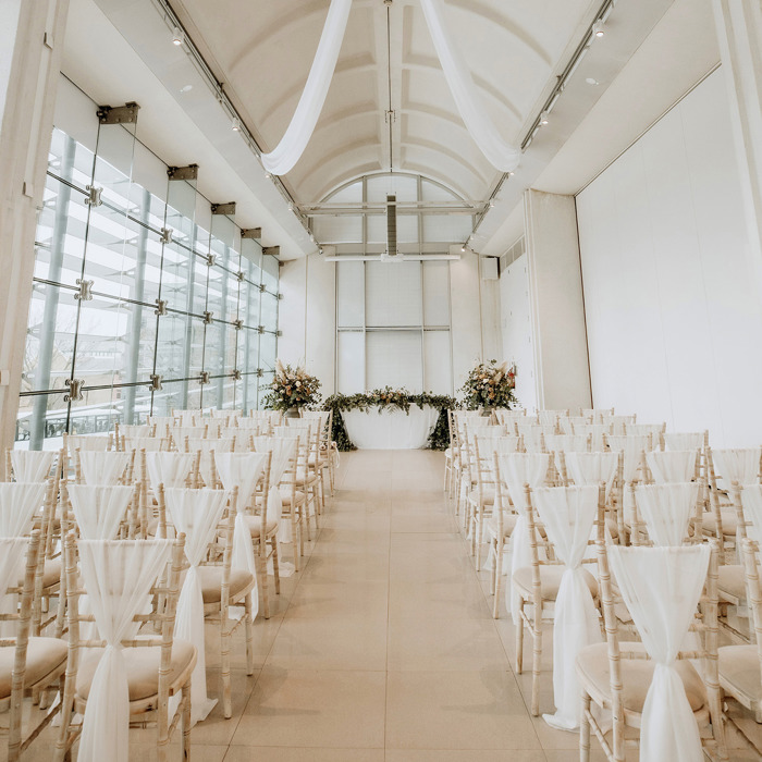 A contemporary white room with a vaulted ceiling and floor to ceiling windows on one side dressed for a wedding. Fabric has been hung from the centre of the ceiling and used to decorate rows of chairs. At far end of the room are three floral arrangements.   