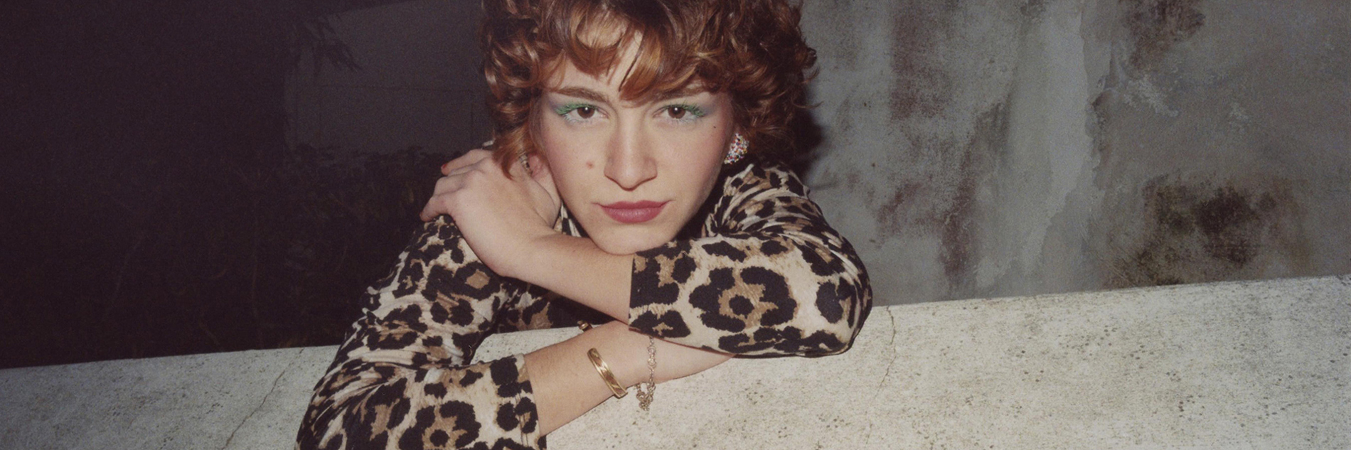 A woman wearing a leopard print dress leans over a wall, looking directly at the camera with a confident gaze. She has green mascara on her eyelashes, red curly hair and wears golden jewellery.