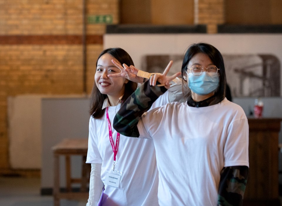Two young people standing in a gallery space - one is wearing a face mask, both are making a peace sign with one hand.  