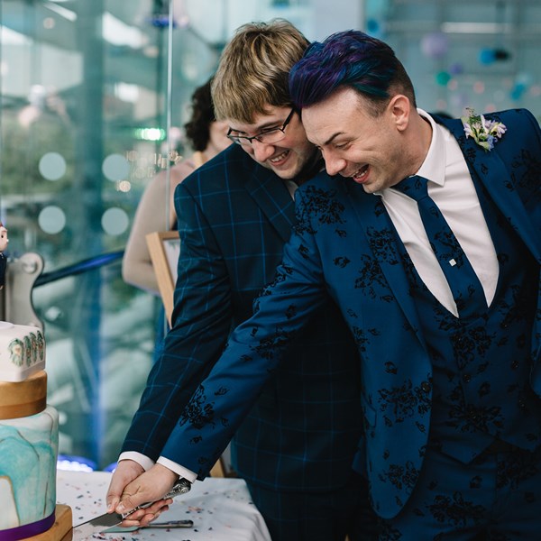 Two grooms in suits hold a knife together to cut a wedding cake.