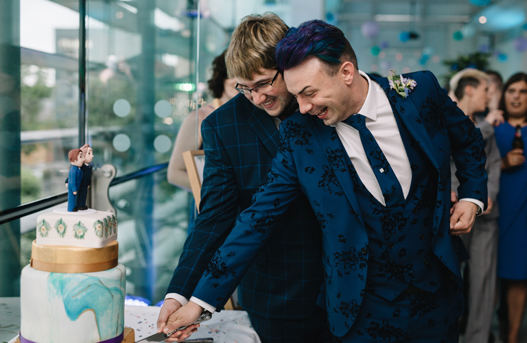 Two grooms in suits hold a knife together to cut a wedding cake.