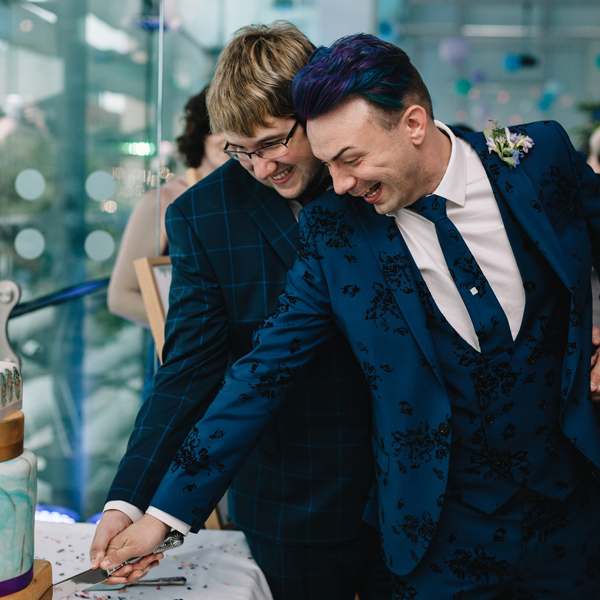 Two grooms in suits hold a knife together to cut a wedding cake.