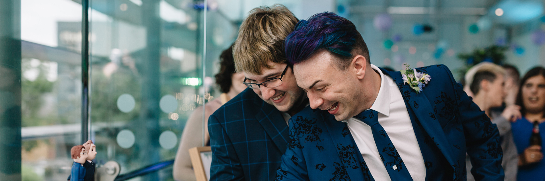 Two grooms in suits hold a knife together to cut a wedding cake.