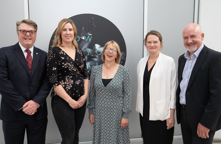 A group of 5 people in business attire standing beside a poster for the exhibition.