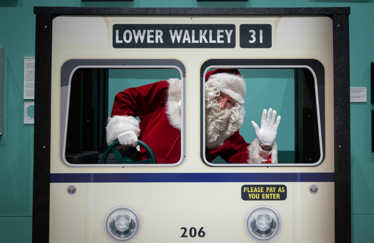 A photograph of Santa pretending to drive the tram in Weston Park Museum, waving at the camera.