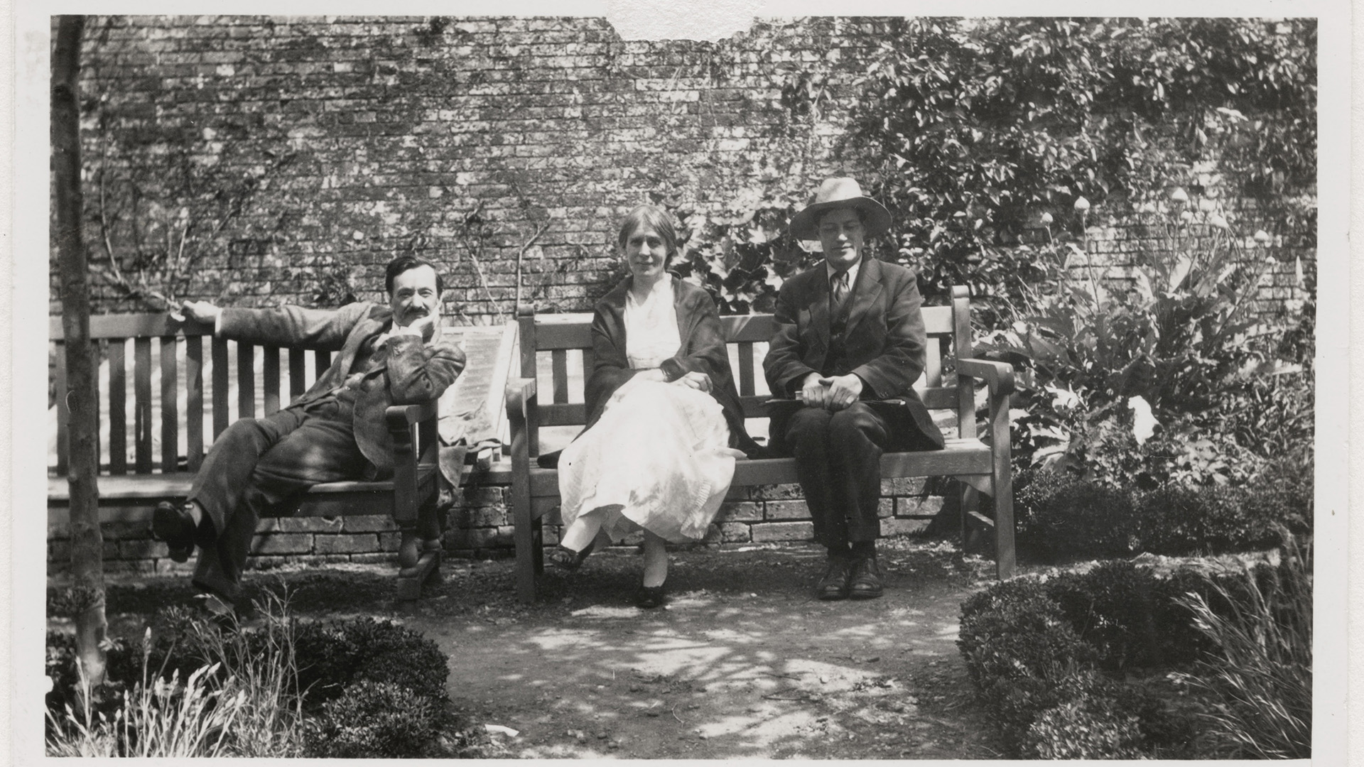An old black and white photograph of three people (one woman, two men) sat on benches in walled garden, sunlight shining from above.
