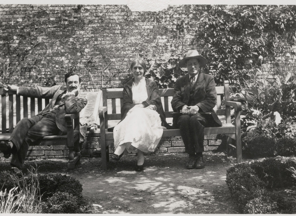 An old black and white photograph of three people (one woman, two men) sat on benches in walled garden, sunlight shining from above.