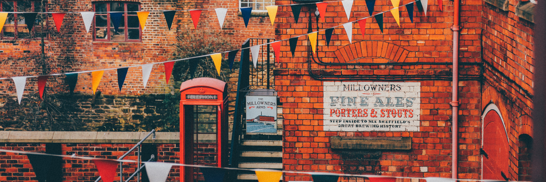 A photograph of industrial buildings, a phone box and a bench. Bunting is hung between the buildings. A sign on the building says "The Millowners Arms" and on the side of the wall there is a large painted sign which says "Millowners fine ales, porters and stouts". 