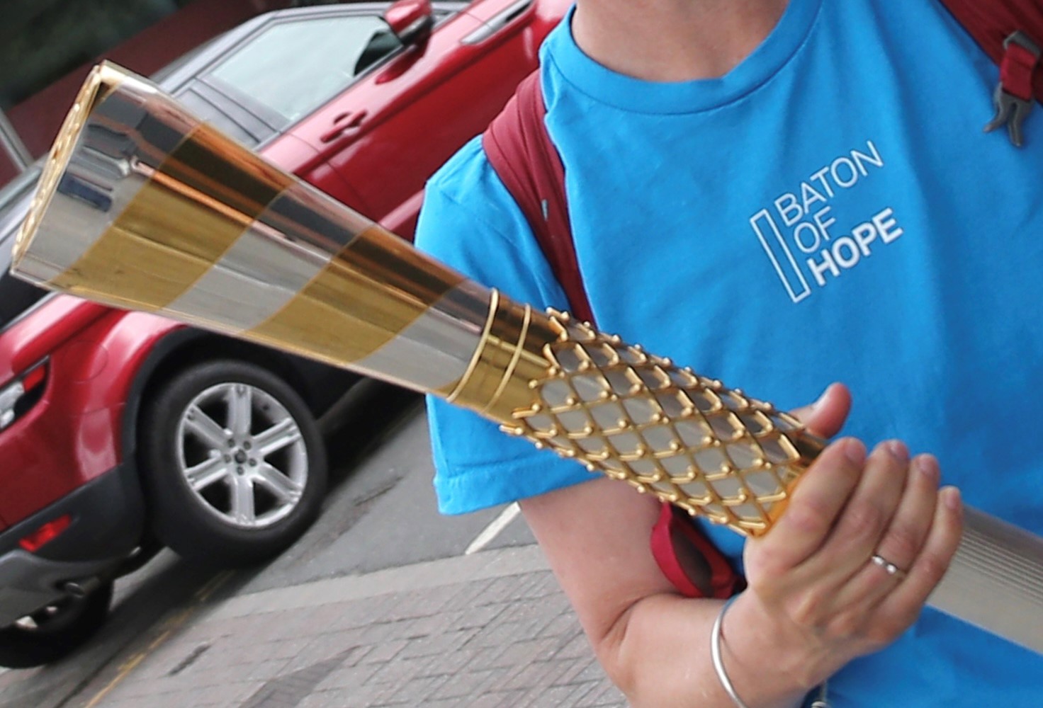 A close-up image of someone in a blue t-shirt holding the baton of hope, a silver and gold baton. The blue shirt has white text which says baton of hope. A red car is visible behind the person.