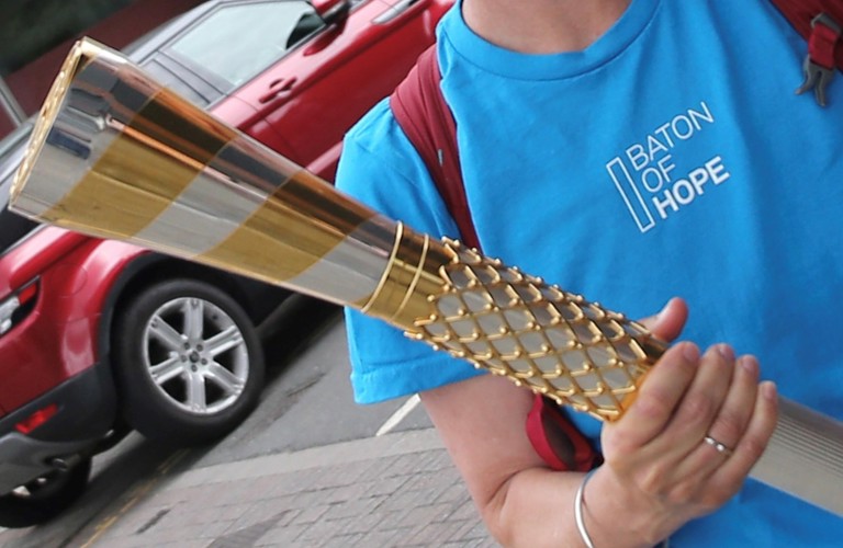 A close-up image of someone in a blue t-shirt holding the baton of hope, a silver and gold baton. The blue shirt has white text which says baton of hope. A red car is visible behind the person.