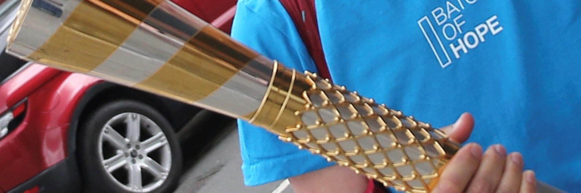 A close-up image of someone in a blue t-shirt holding the baton of hope, a silver and gold baton. The blue shirt has white text which says baton of hope. A red car is visible behind the person.