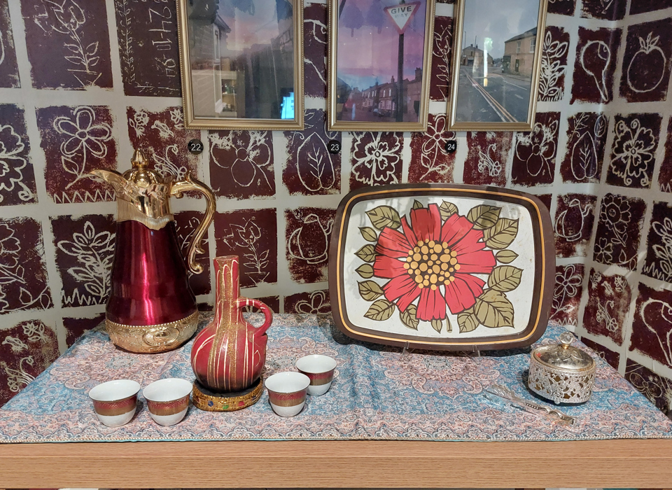 A photograph of a table covered in a paisley print tablecloth, against a wall papered with brown and white floral tiled pattern. On the table, there is a tall red and gold lidded jug, a slightly smaller maroon vessel splattered with white paint. There is also a serving tray with a large red flower painted in the centre. There are also several small, handle-less teacups on the table. 
