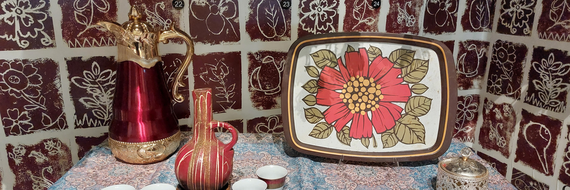A photograph of a table covered in a paisley print tablecloth, against a wall papered with brown and white floral tiled pattern. On the table, there is a tall red and gold lidded jug, a slightly smaller maroon vessel splattered with white paint. There is also a serving tray with a large red flower painted in the centre. There are also several small, handle-less teacups on the table.