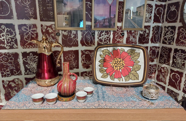 A photograph of a table covered in a paisley print tablecloth, against a wall papered with brown and white floral tiled pattern. On the table, there is a tall red and gold lidded jug, a slightly smaller maroon vessel splattered with white paint. There is also a serving tray with a large red flower painted in the centre. There are also several small, handle-less teacups on the table.