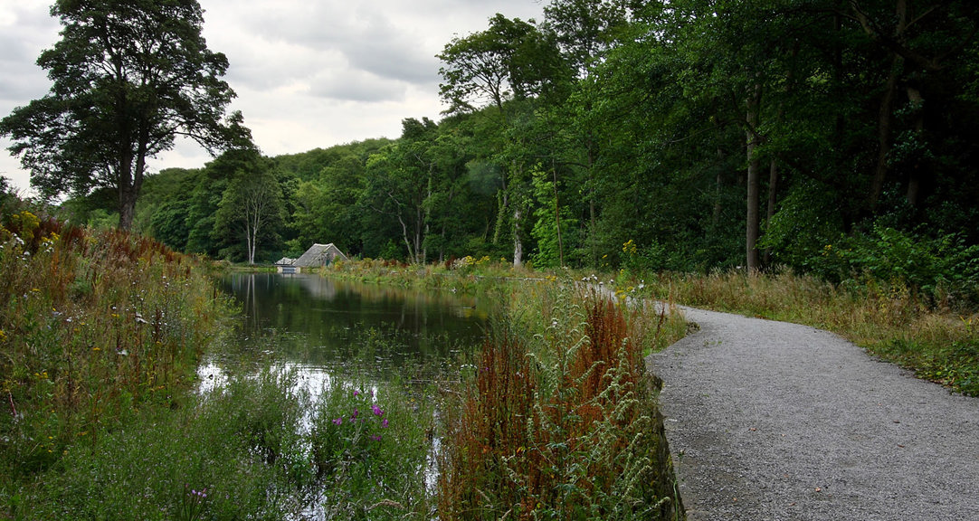 A body of water with a stone path that follows the edge of the water. The path leads to a small building which is surrounded by trees and vegetation.