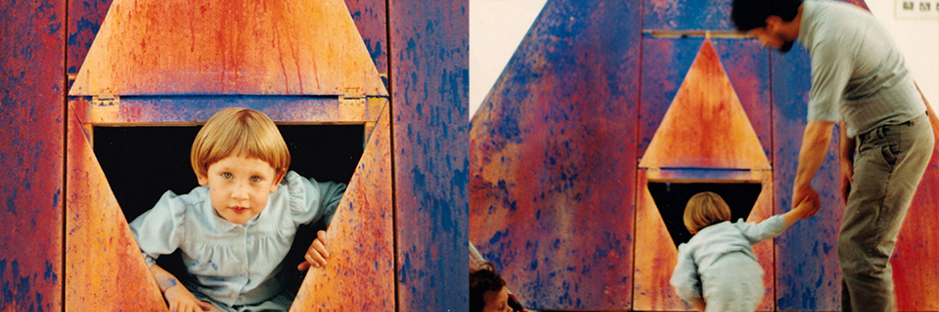 A photograph of a colourful art installation. L-R: a child is looking out from a triangle shaped window; an adult and child are looking into a triangle shaped window.  