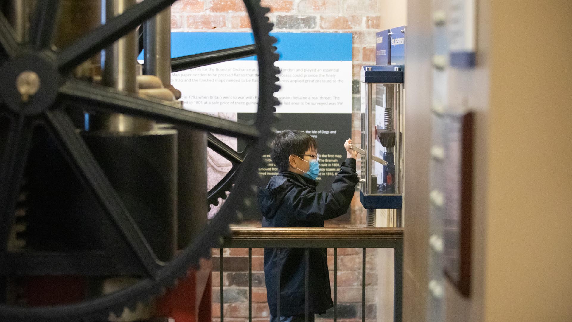 A child wearing a medical facemask operating an interactive scale model of a hydraulic press alongside the real thing.