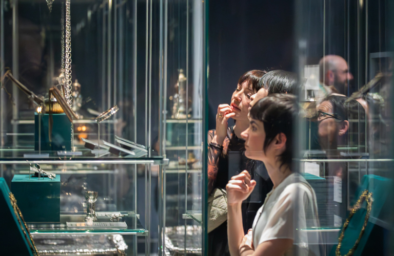 A group of people look at decorative metalwork objects in glass cases in a gallery.