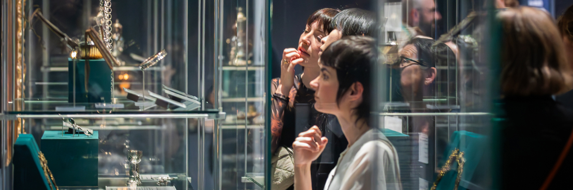 A group of people look at decorative metalwork objects in glass cases in a gallery.