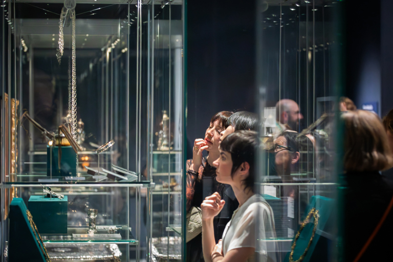 A group of people look at decorative metalwork objects in glass cases in a gallery.