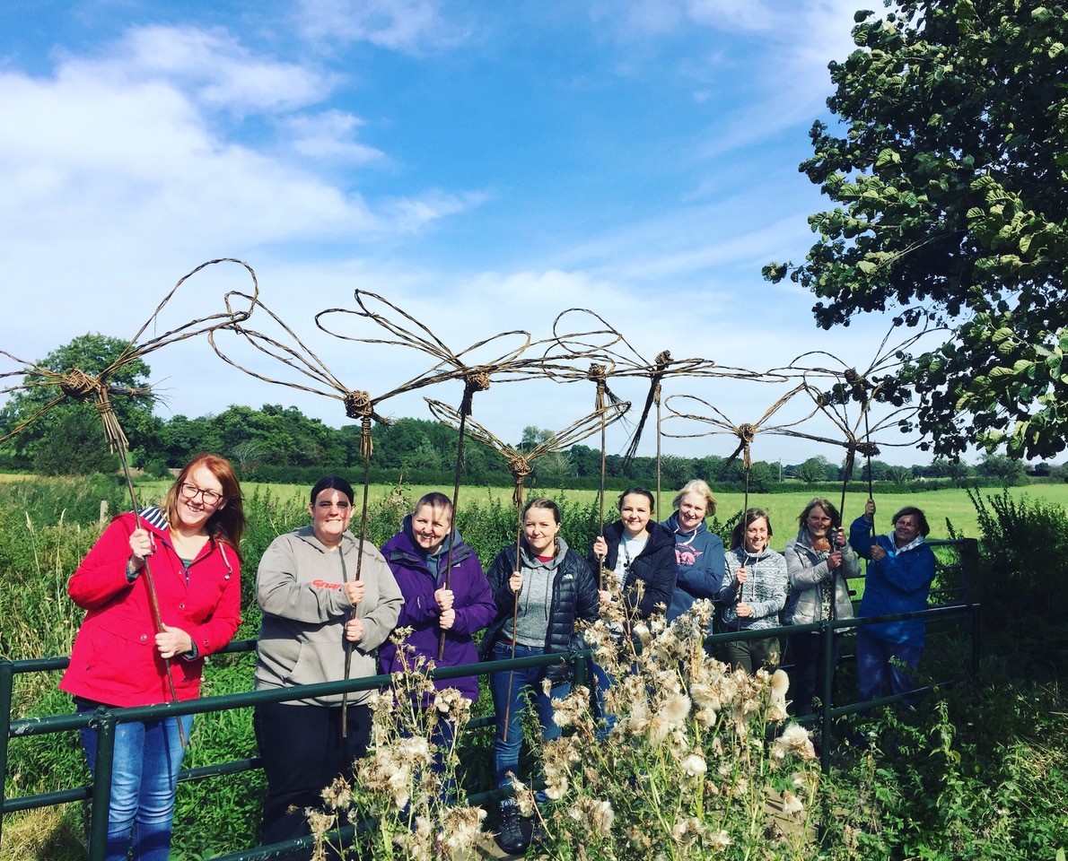Workshop participants standing in a field holding dragonfly sculptures made of willow. The sculptures are very large and stand on sticks above their heads.
