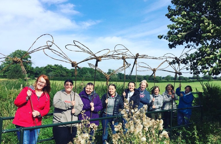 Workshop participants standing in a field holding dragonfly sculptures made of willow. The sculptures are very large and stand on sticks above their heads.