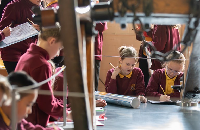 A group of school children writing on clipboards among historic industrial machinery.