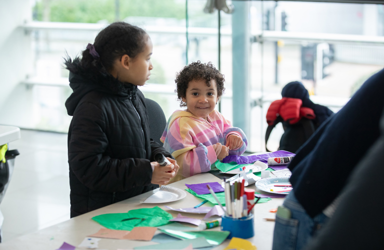 Two children at a table full of crafting materials, including tissue paper, cutting and sticking onto a paper plate.