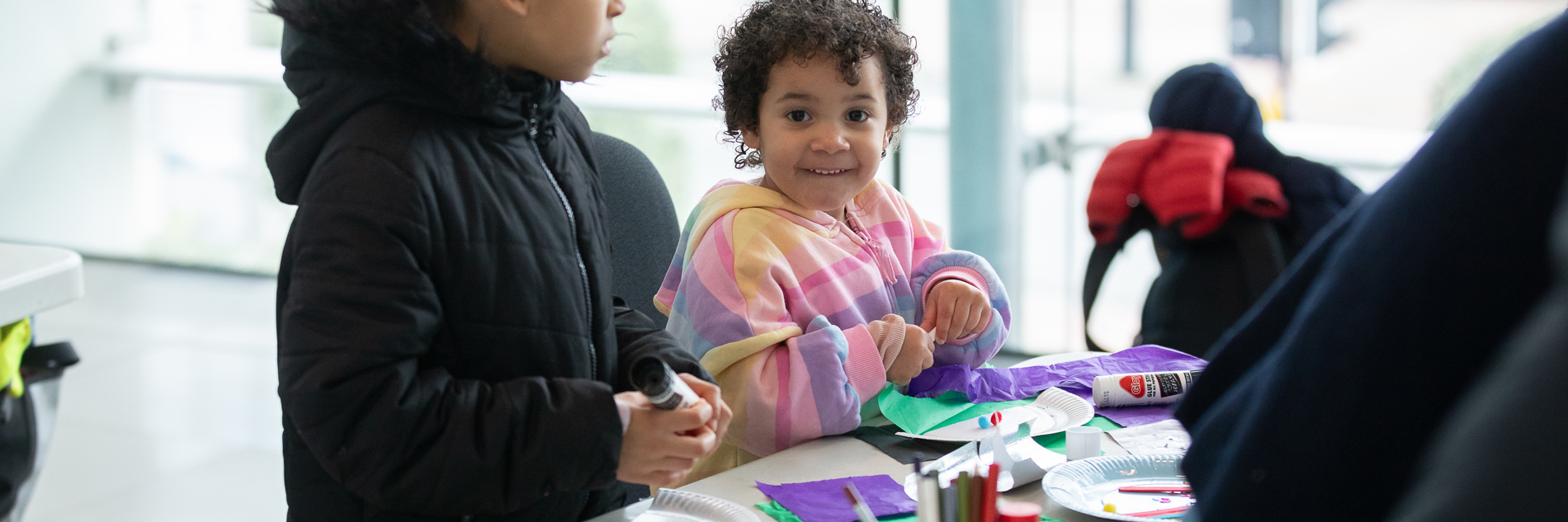 Two children at a table full of crafting materials, including tissue paper, cutting and sticking onto a paper plate.