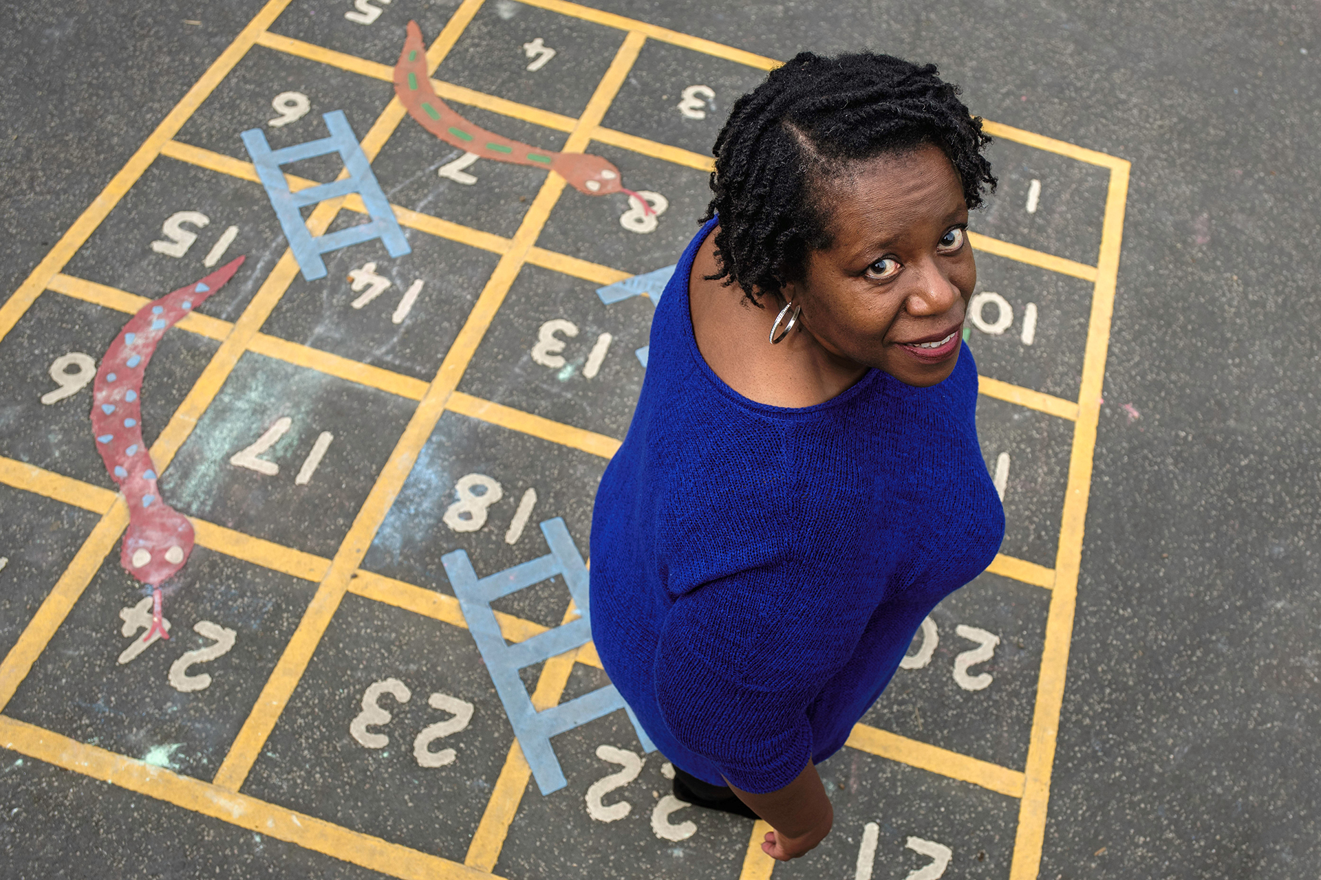 A photograph of a black woman wearing a knitted blue top, looking up at the camera directly into the lens. She is smiling and her silver hoop earring catches the light. The photo is taken from above, so we can see the woman is standing on a school playground game of snakes and ladders, painted directly onto the ground. 