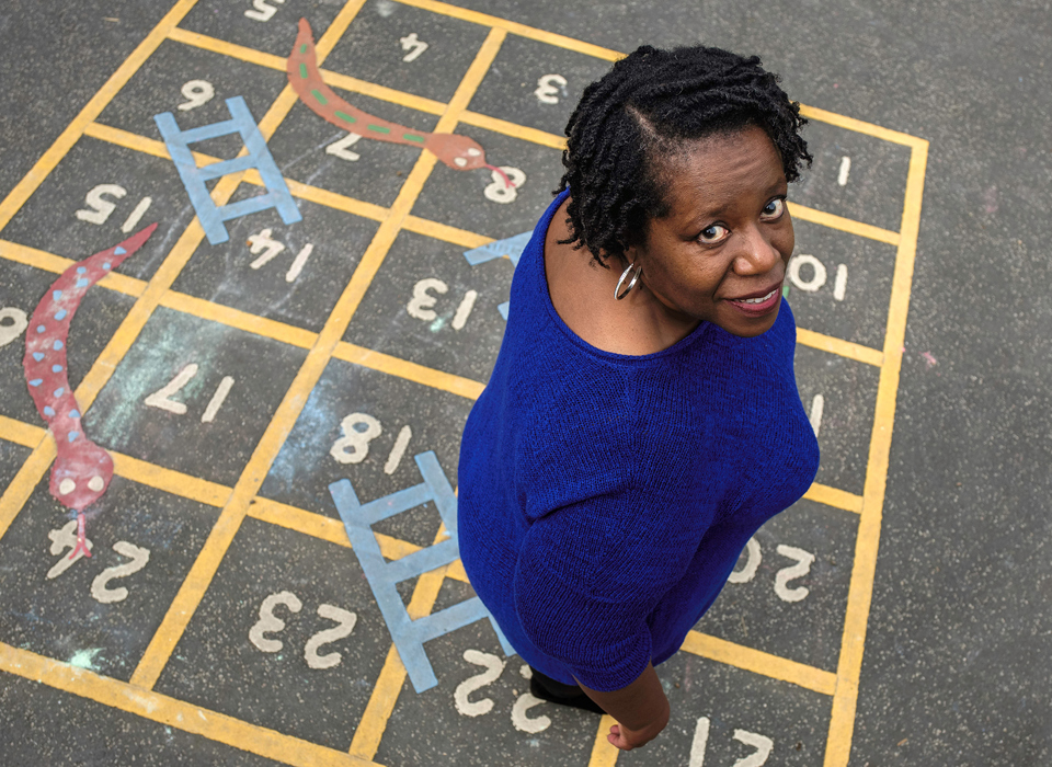 A photograph of a black woman wearing a knitted blue top, looking up at the camera directly into the lens. She is smiling and her silver hoop earring catches the light. The photo is taken from above, so we can see the woman is standing on a school playground game of snakes and ladders, painted directly onto the ground. 