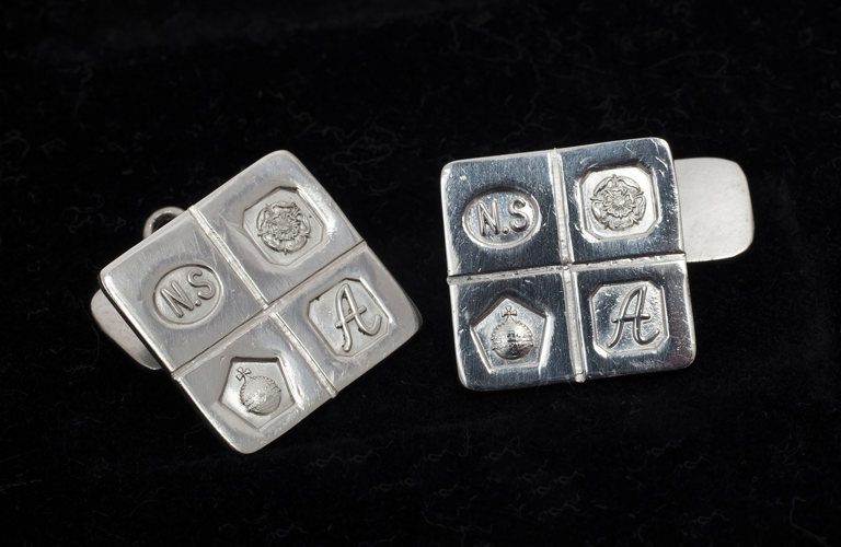 A pair of silver square cufflinks embossed with 4 symbols - the letters N.S, a flower, an orb and the letter A.  