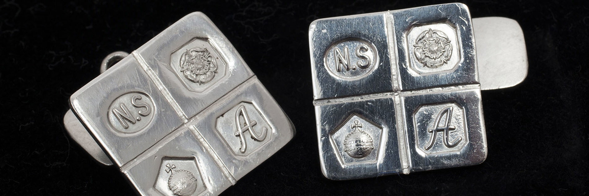 A pair of silver square cufflinks embossed with 4 symbols - the letters N.S, a flower, an orb and the letter A.  