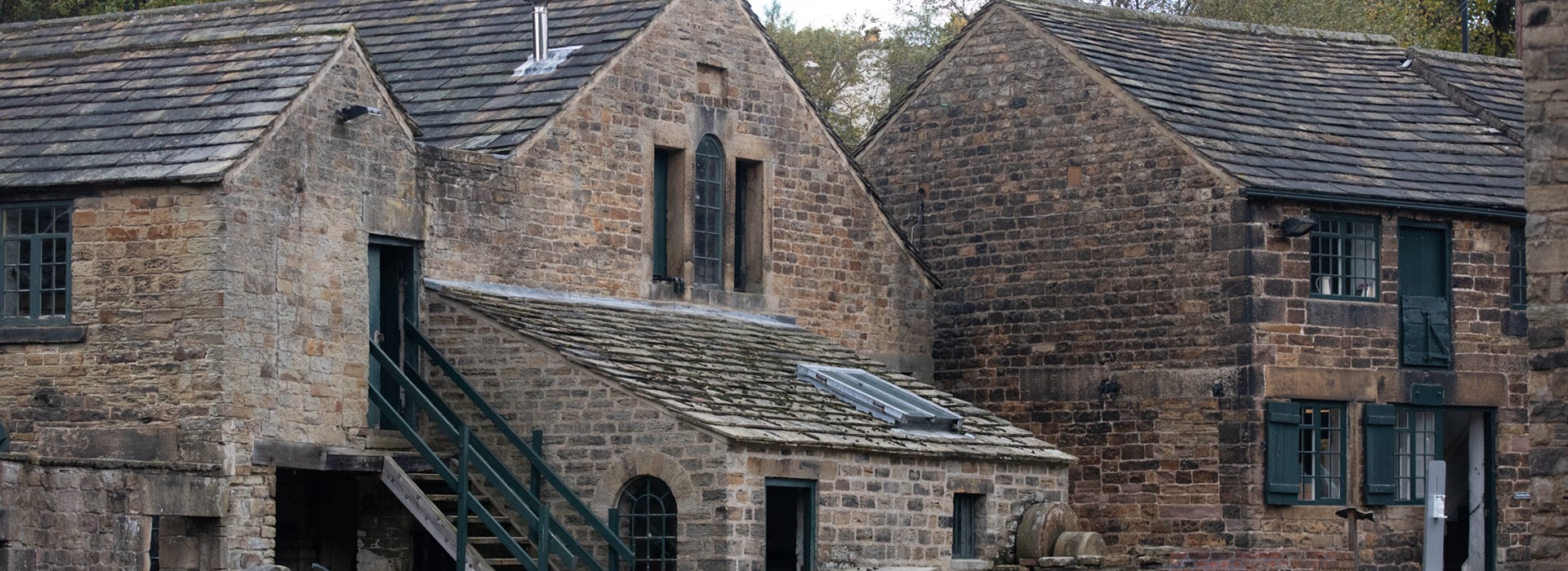 A collection of historic industrial stone buildings. They have large green windows and the building to the left has steps leading up to the second storey. There are trees behind.